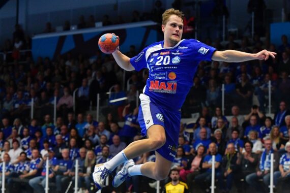 Ein Handballspieler in einem blauen Trikot springt in der Luft, um den Ball während eines Spiels zu werfen, während im Hintergrund eine große Menge von Zuschauern von der Tribüne aus zusieht.
