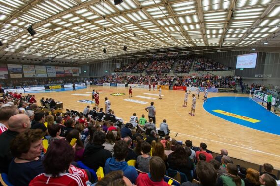 Eine überfüllte Halle mit Zuschauern, die ein Handballspiel verfolgen. Spieler in rot-weißen Trikots stehen auf dem Spielfeld, und die Wände sind mit Bannern und Werbung geschmückt. Helle Lichter erhellen den Raum.