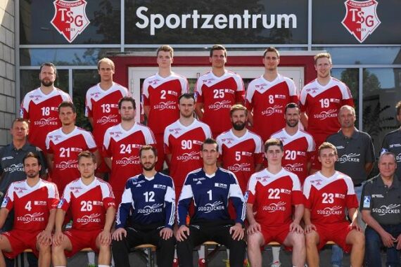 Eine Herren-Handballmannschaft in rot-weißen Trikots posiert für ein Gruppenfoto mit Trainern und Mitarbeitern vor einem Gebäude mit der Aufschrift Sportzentrum. Drei Reihen: sitzendes Personal, mittlere Reihe stehend und hintere Reihe auf einer Stufe stehend.