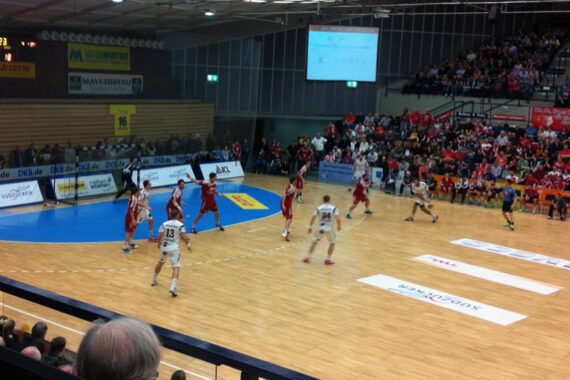 Ein Handballspiel wird in einer Halle ausgetragen, die Mannschaften tragen rote und weiße Trikots. Das Spielfeld ist von Zuschauern auf der Tribüne umgeben, und auf der blau-hellbraunen Spielfläche ist Werbung zu sehen.