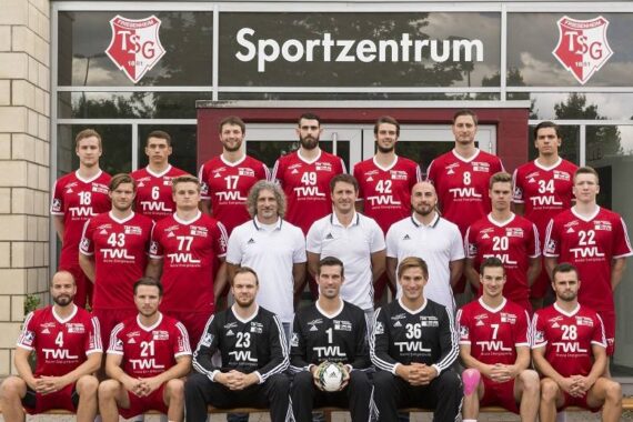 Eine Männerhandballmannschaft in roten und schwarzen Trikots stellt sich für ein Gruppenfoto vor einem Gebäude mit der Aufschrift Sportzentrum auf. Vier Trainer oder Betreuer in weißen Hemden stehen in der mittleren Reihe. Zwei Torhüter in schwarzen Hemden sitzen in der vorderen Reihe.