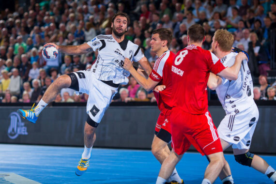 Ein Handballspieler in einem weißen Trikot springt, um den Ball zu schießen, während drei gegnerische Spieler in Rot versuchen, ihn während eines Spiels zu blockieren, während im Hintergrund ein dicht gedrängtes Publikum zusieht.