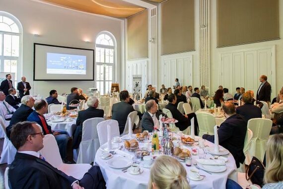 Eine Gruppe von Personen in Geschäftskleidung nimmt an einer formellen Sitzung oder Konferenz in einem hellen, eleganten Raum mit hohen Decken teil; die Tische sind mit Speisen und Getränken gedeckt, und auf einem großen Bildschirm wird eine Präsentation gezeigt.