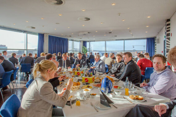 In einem hellen, modernen Raum mit großen Fenstern und Blick auf die Stadt sitzt eine Gruppe von Menschen, meist in Geschäftskleidung, um einen langen Tisch mit Frühstücks- oder Brunchgerichten. Einige essen, andere unterhalten sich.