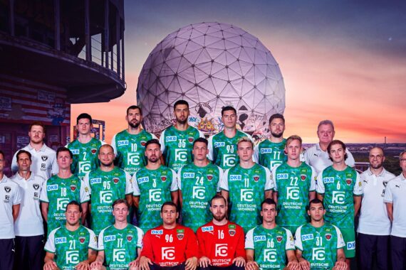 Eine Herren-Handballmannschaft in grün-weißen Trikots posiert für ein Gruppenfoto im Freien bei Sonnenuntergang, mit Trainern in weißen Hemden und einem großen geodätischen Kuppelbau im Hintergrund.