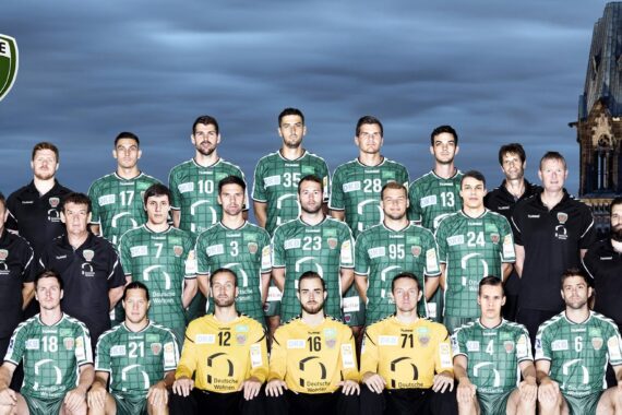 Eine Handballmannschaft in grünen Trikots und gelben Torwarttrikots posiert in einer Reihe mit Trainern vor einer Stadtkulisse mit einem historischen Gebäude. In der linken oberen Ecke ist das Logo der Füchse Berlin zu sehen.