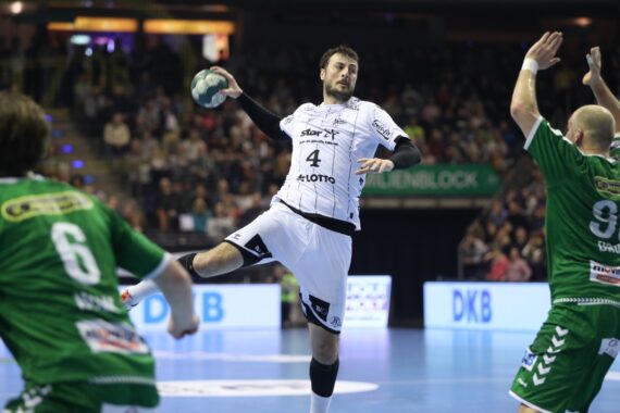 Ein Handballspieler in einem weißen Trikot springt, um den Ball zu werfen, während zwei Spieler in grünen Trikots versuchen, ihn während eines Spiels in einer mit Zuschauern gefüllten Halle zu blockieren.