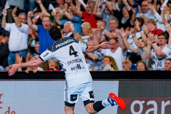 Ein Handballspieler, der ein weißes Trikot mit der Nummer 4 trägt, jubelt mit ausgestreckten Armen und läuft auf jubelnde Fans in einer überfüllten Arena zu. Das Publikum wirkt aufgeregt und jubelnd.