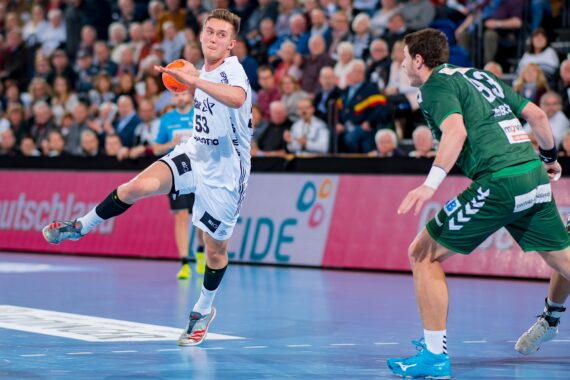 Ein Handballspieler in einem weißen Trikot springt und bereitet sich auf den Wurf vor, während ein Spieler in einem grünen Trikot den Ball abwehrt. Das Spiel findet in einer Halle statt und wird von Zuschauern auf der Tribüne verfolgt.