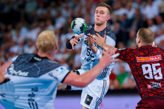 Drei männliche Handballspieler kämpfen während eines Spiels um den Ball, wobei zwei Spieler weiße Trikots und einer ein rotes Trikot trägt. Der Spieler in der Mitte fängt oder wirft den Ball in der Luft. Im Hintergrund sieht eine Menschenmenge zu.
