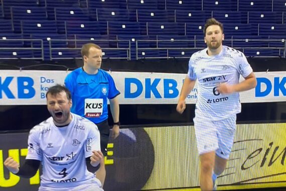 Zwei männliche Handballspieler in weißen Trikots feiern leidenschaftlich auf dem Spielfeld, der eine kniend und mit geballten Fäusten schreiend. Ein Schiedsrichter steht hinter ihnen. Im Hintergrund sind leere Plätze in der Arena und ein DKB-Sponsorenbanner zu sehen.
