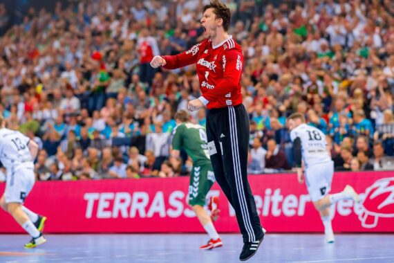 Ein Handballtorwart in einem roten Trikot springt jubelnd über das Spielfeld, während sich andere Spieler in weißen und grünen Trikots im Hintergrund bewegen. Die Arena ist mit einer jubelnden Menge gefüllt.