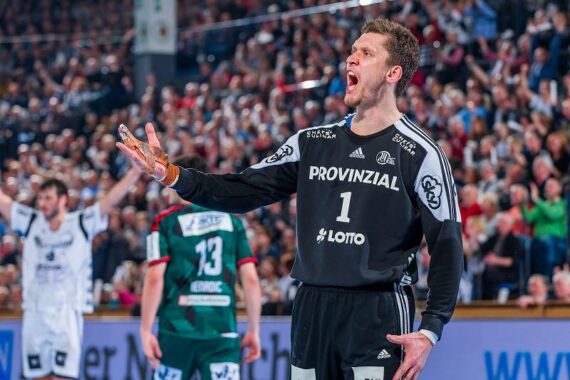 Ein männlicher Handballtorwart in einem schwarzen Trikot mit der Aufschrift PROVINZIAL und der Nummer 1 ruft während eines Spiels und gestikuliert mit seiner rechten Hand. Im Hintergrund sind unscharfe Spieler und eine jubelnde Menge zu sehen.