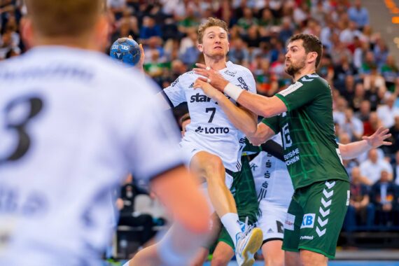Ein männlicher Handballspieler in einem weißen Trikot springt, um den Ball zu schießen, während ein Gegner in einem grünen Trikot ihn blockiert. Ein anderer Spieler in weißem Trikot ist im Vordergrund zu sehen. Das Spiel findet in einer Halle statt und wird von einer Zuschauermenge verfolgt.