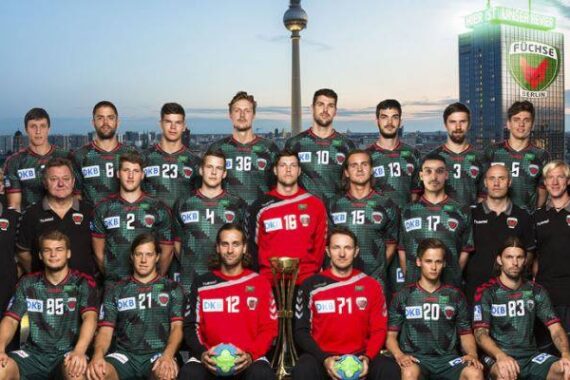 Eine Männer-Handballmannschaft in grün-schwarzen Trikots posiert für ein Gruppenfoto im Freien mit ihren Trainern. Im Hintergrund sind die Berliner Skyline und der Fernsehturm zu sehen, sowie ein Gebäude mit dem Logo der Füchse Berlin.