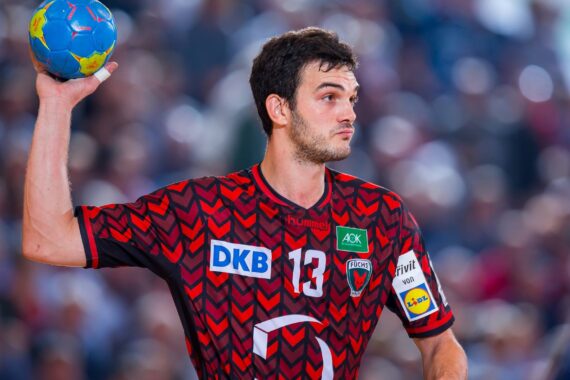 Ein männlicher Handballspieler in einem rot-schwarz gemusterten Trikot bereitet sich während eines Spiels auf den Wurf eines blauen Balls vor, mit einer unscharfen Menschenmenge im Hintergrund.
