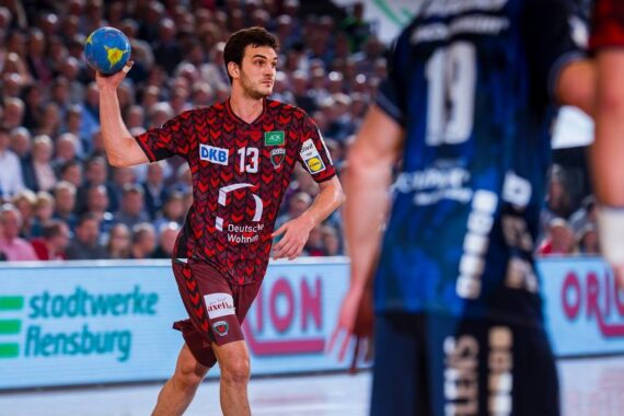 Ein männlicher Handballspieler in einem rot gemusterten Trikot bereitet sich darauf vor, während eines Spiels einen blauen Ball zu werfen, während ein gegnerischer Spieler in einem blauen Trikot im Vordergrund und eine Menschenmenge im Hintergrund zu sehen sind.