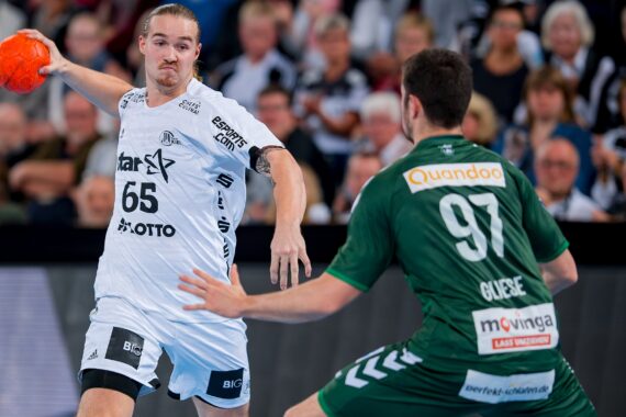 Zwei männliche Handballspieler sind während eines Spiels in Aktion. Der Spieler in Weiß hält einen orangefarbenen Ball und bereitet sich auf den Wurf vor, während der Spieler in Grün verteidigt. Im Hintergrund sind Zuschauer zu sehen.