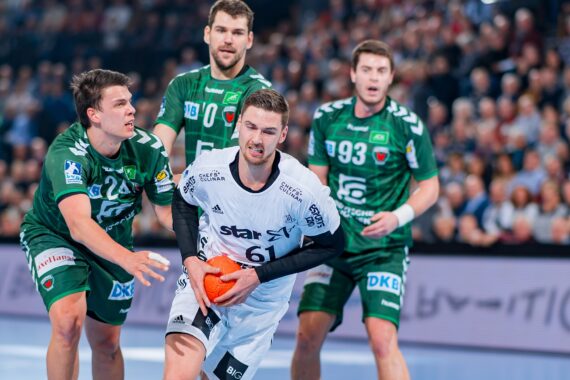 Ein Handballspieler in einem weißen Trikot hält den Ball, während er während eines Spiels von drei Spielern in grünen Trikots verteidigt wird, während im Hintergrund eine Menschenmenge zu sehen ist.