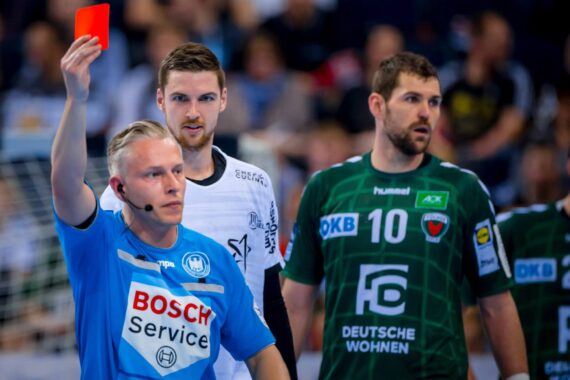 Ein Schiedsrichter in einem blauen Hemd hält während eines Handballspiels eine rote Karte hoch, während zwei männliche Spieler in grün-weißen Trikots zuschauen. Die Szene ist angespannt, mit einer verschwommenen Menge im Hintergrund.