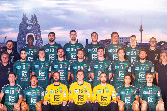Eine Handballmannschaft in grün-schwarzen Trikots posiert für ein Gruppenfoto mit den Trainern vor einer Stadtkulisse mit markanten Skulpturen und einem Fernsehturm.