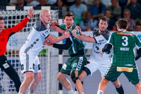 Zwei Spieler in weißen Trikots verteidigen während eines Handballspiels gegen einen Spieler in einem grünen Trikot, während der Torwart in einem orangefarbenen Trikot vom Torraum aus zusieht. Die Szene ist intensiv und actionreich.