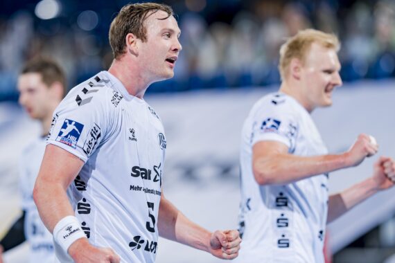 Zwei männliche Handballspieler in weißen Trikots feiern während eines Spiels. Der eine ist im Fokus und sieht entschlossen aus, während der andere im Hintergrund verschwommen ist. Beide haben die Fäuste geballt, und hinter ihnen ist eine Menschenmenge zu sehen.