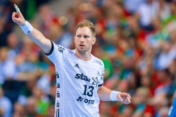 Ein männlicher Handballspieler in einem weißen Trikot mit der Nummer 13 zeigt mit der rechten Hand während eines Spiels auf eine unscharfe Menschenmenge im Hintergrund.