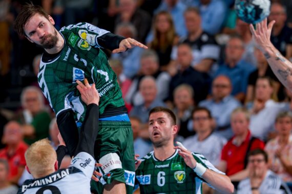 Ein männlicher Handballspieler in einem grünen Trikot springt, um den Ball zu werfen, während er von Gegnern in weißen Trikots verteidigt wird, während die Zuschauer im Hintergrund zuschauen.