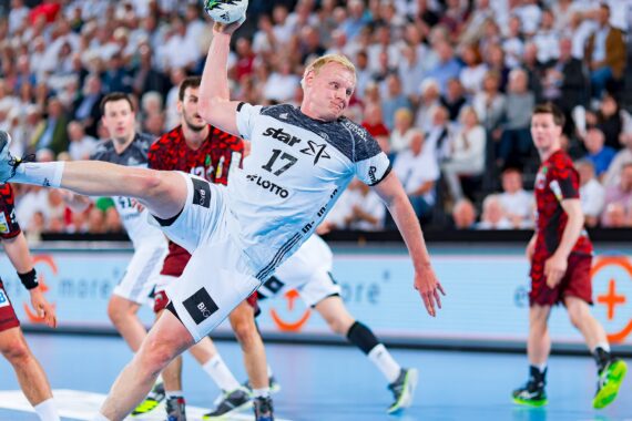 Ein Handballspieler in einem weißen Trikot, in der Luft und im Begriff, den Ball zu werfen, ist von Spielern in roten Trikots auf einem Hallenplatz umgeben, während im Hintergrund eine große Menschenmenge zusieht.
