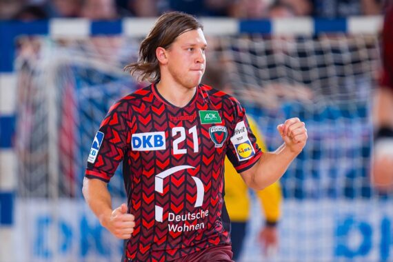Ein männlicher Handballspieler in einem rot gemusterten Trikot mit der Nummer 21 läuft auf dem Spielfeld, mit einem unscharfen Tor und einem Schiedsrichter im Hintergrund.