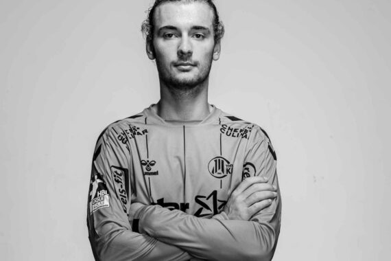 Ein junger Mann mit langem, nach hinten gebundenem Haar, der ein mit Sponsorenlogos versehenes Sporttrikot trägt, steht mit verschränkten Armen und blickt selbstbewusst in die Kamera vor einem schlichten Hintergrund. Das Bild ist in schwarz-weiß gehalten.