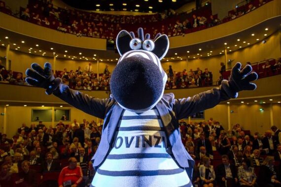 Eine Person in einem Zebra-Maskottchenkostüm steht mit weit ausgebreiteten Armen auf einer Theaterbühne und blickt auf ein großes, sitzendes Publikum. Auf dem Hemd des Zebras steht OVINZI, und die Bühnenbeleuchtung erhellt die Szene.