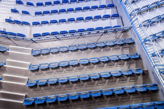 Reihen leerer blauer Stadionsitze von oben gesehen, mit Metallgeländern und Treppen, die die Abschnitte voneinander trennen. Die Beleuchtung wirft Schatten, und die Sitze sind in einem Muster auf dem Betonboden angeordnet.