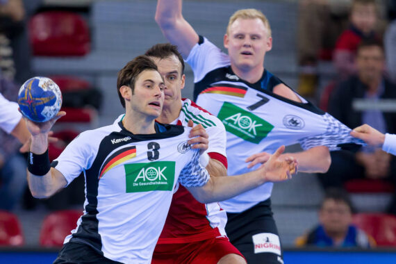 Zwei männliche Handballspieler in weißen und schwarzen Trikots mit den Farben und Logos der deutschen Flagge kämpfen auf einem Hallenplatz um den Ball gegen einen Spieler in einem rot-weißen Trikot, während im Hintergrund Zuschauer sitzen.