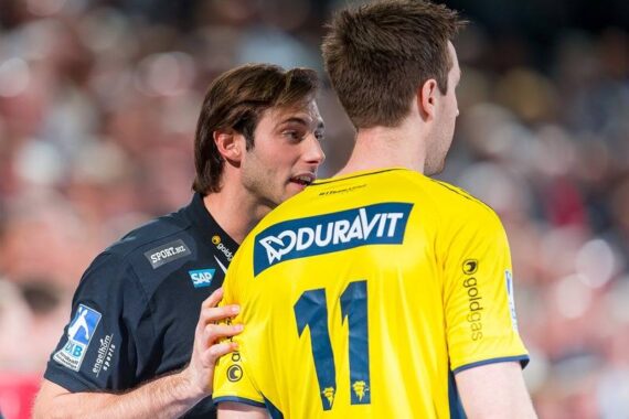 Ein Trainer in einem dunklen Hemd spricht mit einem Handballspieler, der ein gelbes Trikot mit der Nummer 11 und DURAVIT auf dem Rücken trägt, während eines Spiels. Der Hintergrund ist unscharf, eine Menschenmenge ist zu sehen.