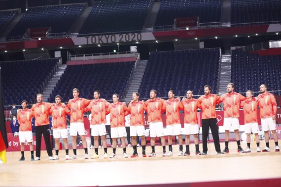 Eine Handballmannschaft der Männer in roten Hemden und weißen Shorts steht in einer Halle in einer Reihe und legt sich gegenseitig die Arme um die Schultern, wahrscheinlich vor einem Spiel bei den Olympischen Spielen 2020 in Tokio. Die Tribünen sind größtenteils leer.