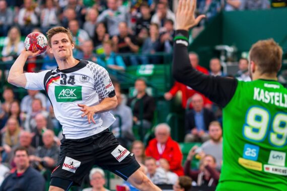 Ein männlicher Handballspieler in einem weiß-schwarzen Trikot bereitet sich darauf vor, den Ball zu werfen, während ein Torwart in einem grünen Trikot mit KASTELIC 99 das Tor blockiert. Die Zuschauer auf der Tribüne verfolgen das intensive Spiel.