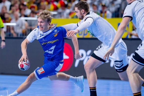 Ein Handballspieler in einem blauen Trikot läuft mit dem Ball in der Hand, dicht verfolgt und verteidigt von einem Gegner in einem weißen Trikot während eines Hallenspiels. Im Hintergrund sind Zuschauer und Schiedsrichter zu sehen.