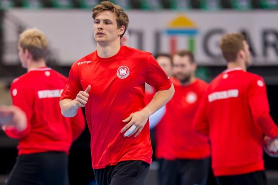 Ein junger männlicher Sportler in einem roten Hemd joggt in der Halle und hält einen Handball in der Hand, während andere, ähnlich gekleidete Mannschaftskameraden im Hintergrund verschwommen zu sehen sind.