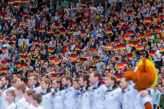 Eine große Menge begeisterter Fans, von denen viele kleine deutsche Fahnen schwenken, füllt die Plätze im Stadion. Im Vordergrund steht eine Reihe von Sportlern in weißen Trikots im Mittelpunkt, unter denen ein Maskottchen zu sehen ist.