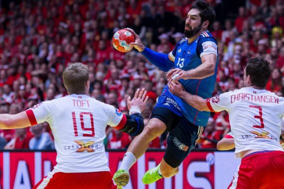 Ein Handballspieler in einem blauen Trikot springt zum Schuss, während zwei Verteidiger in weißen und roten Trikots versuchen, ihn abzuwehren. Die Zuschauer im Hintergrund sind rot gekleidet.