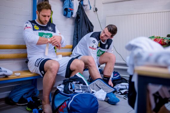 Zwei männliche Sportler in Sportuniformen sitzen auf einer Bank in der Umkleidekabine. Einer bandagiert sein Knie, während der andere zusieht. Sporttaschen und Kleidung liegen verstreut auf dem Boden, und an der Wand hinter ihnen hängen Mannschaftsutensilien.