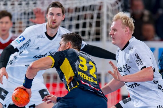 Drei männliche Handballspieler sind während eines Spiels in Aktion. Zwei Spieler in Weiß verteidigen gegen einen Spieler in einem dunklen Trikot, der einen orangefarbenen Ball hält. Im Hintergrund sind ein Netz und eine unscharfe Zuschauermenge zu sehen.