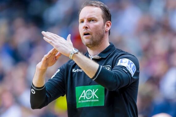 Ein Mann in einer schwarzen Sportuniform gestikuliert mit den Händen und scheint während einer Indoor-Sportveranstaltung Anweisungen zu geben. Der Hintergrund ist mit Zuschauern unscharf.