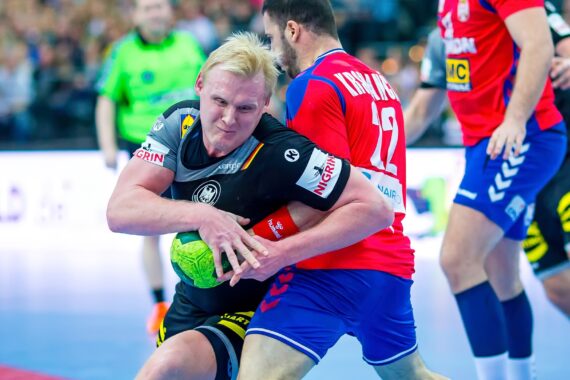 Ein Handballspieler in einem schwarzen Trikot hält den grünen Ball fest umklammert, während er von einem Gegner in einem roten Trikot verteidigt wird, während andere Spieler und ein Schiedsrichter während eines intensiven Spiels zusehen.