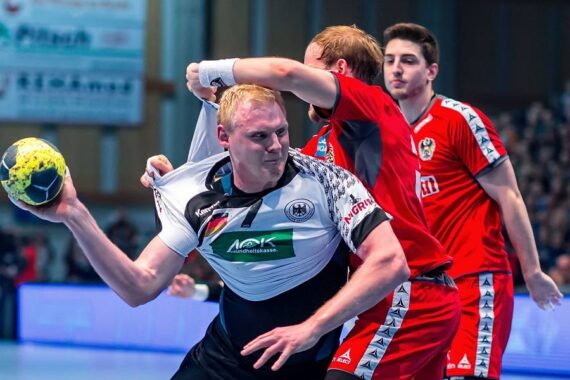 Ein männlicher Handballspieler in einem weißen Trikot wird von einem Gegner in einem roten Trikot gepackt und zurückgehalten, als er versucht, den Ball zu werfen, während ein anderer Spieler in einem roten Trikot in der Nähe zusieht.