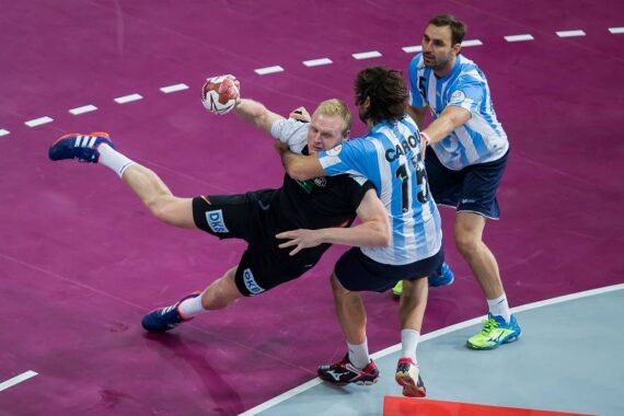 Ein dynamisches Handballspiel zeigt einen Spieler in Schwarz, der in der Luft springt, um zu schießen, während zwei Spieler in Blau und Weiß versuchen, ihn auf einem lilafarbenen Feld zu blockieren.