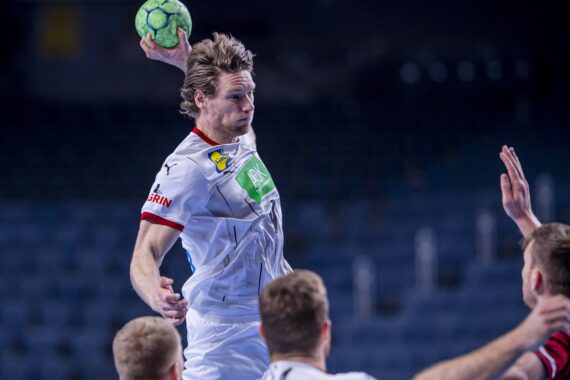 Ein männlicher Handballspieler in einem weißen Trikot springt, um einen grünen Ball zu werfen, während drei gegnerische Spieler in dunklen Trikots versuchen, ihn in einer Halle zu blockieren.