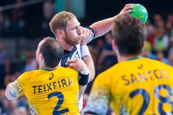 Ein Handballspieler in einem schwarz-weißen Trikot springt, um einen grünen Ball zu werfen, während zwei Verteidiger in gelben Trikots, die mit Teixeira (2) und Santos (28) bezeichnet sind, versuchen, ihn während eines Spiels zu blockieren.
